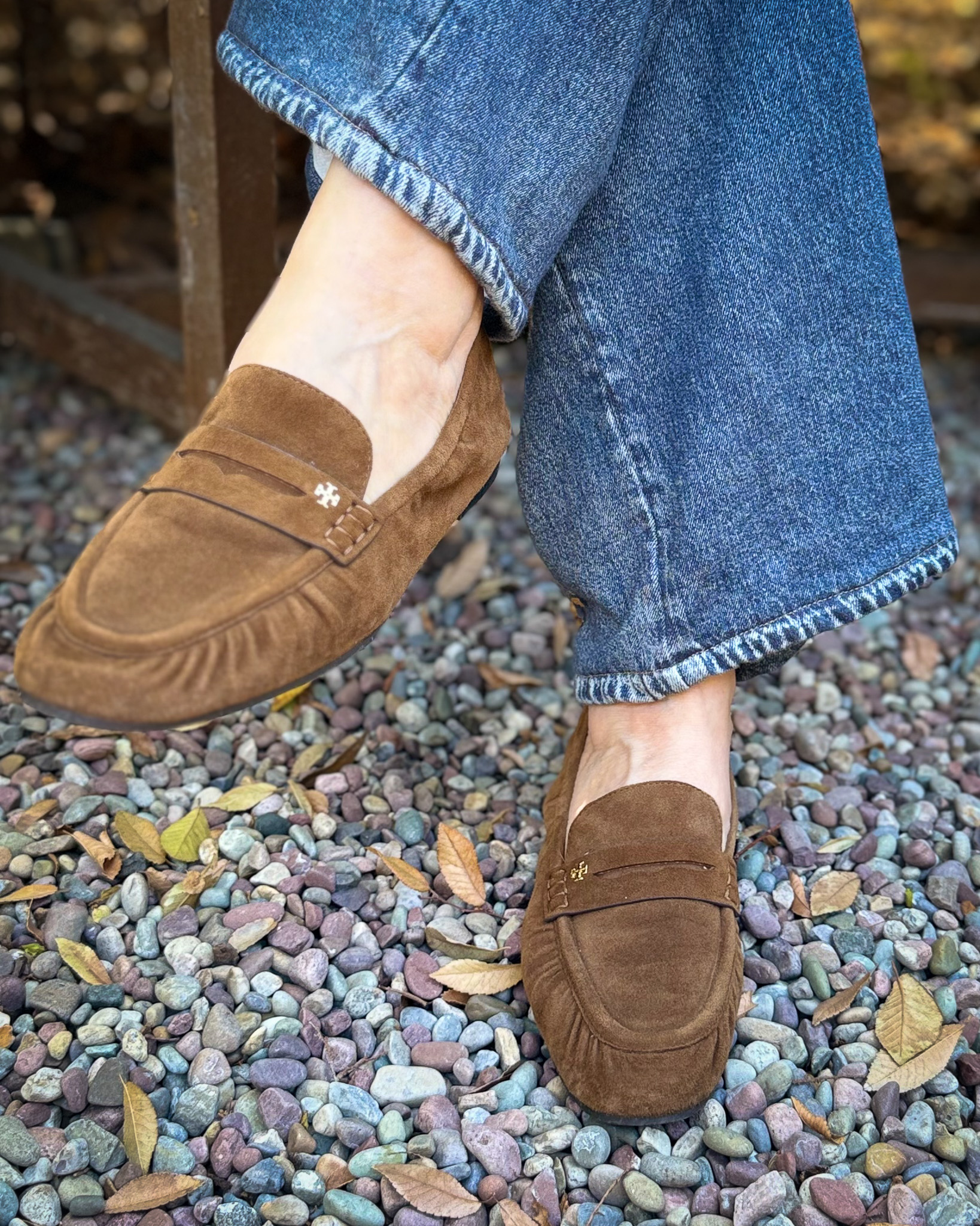 Cognac suede Tory Burch Kira loafers worn with medium-wash wide-leg jeans, photographed on a pebbled path.