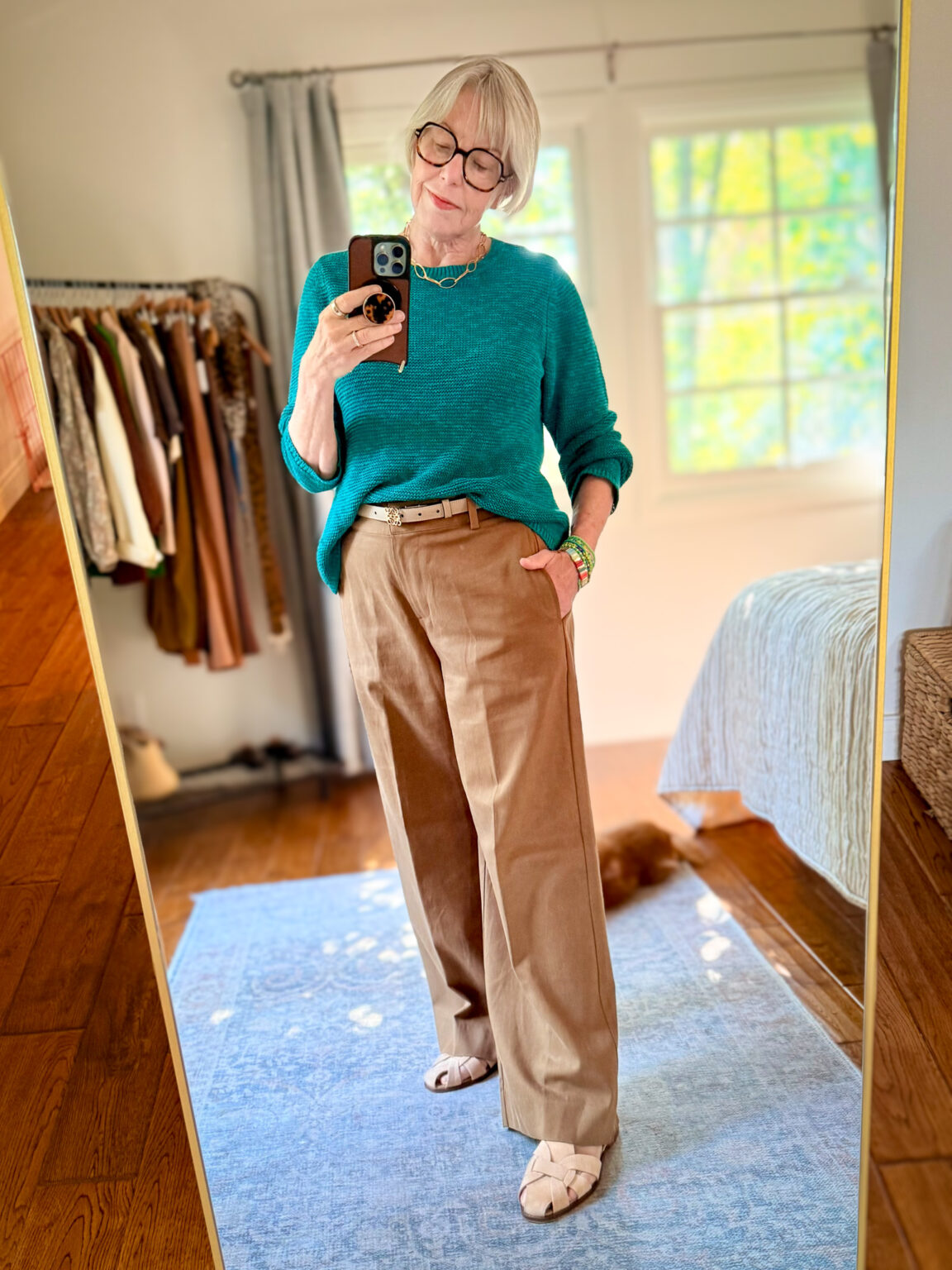 Mirror selfie of a woman wearing a teal knit sweater tucked into wide-leg tan trousers, with a tan belt, woven flat sandals, gold jewelry, and a beaded bracelet, standing on a light blue rug in a bedroom.