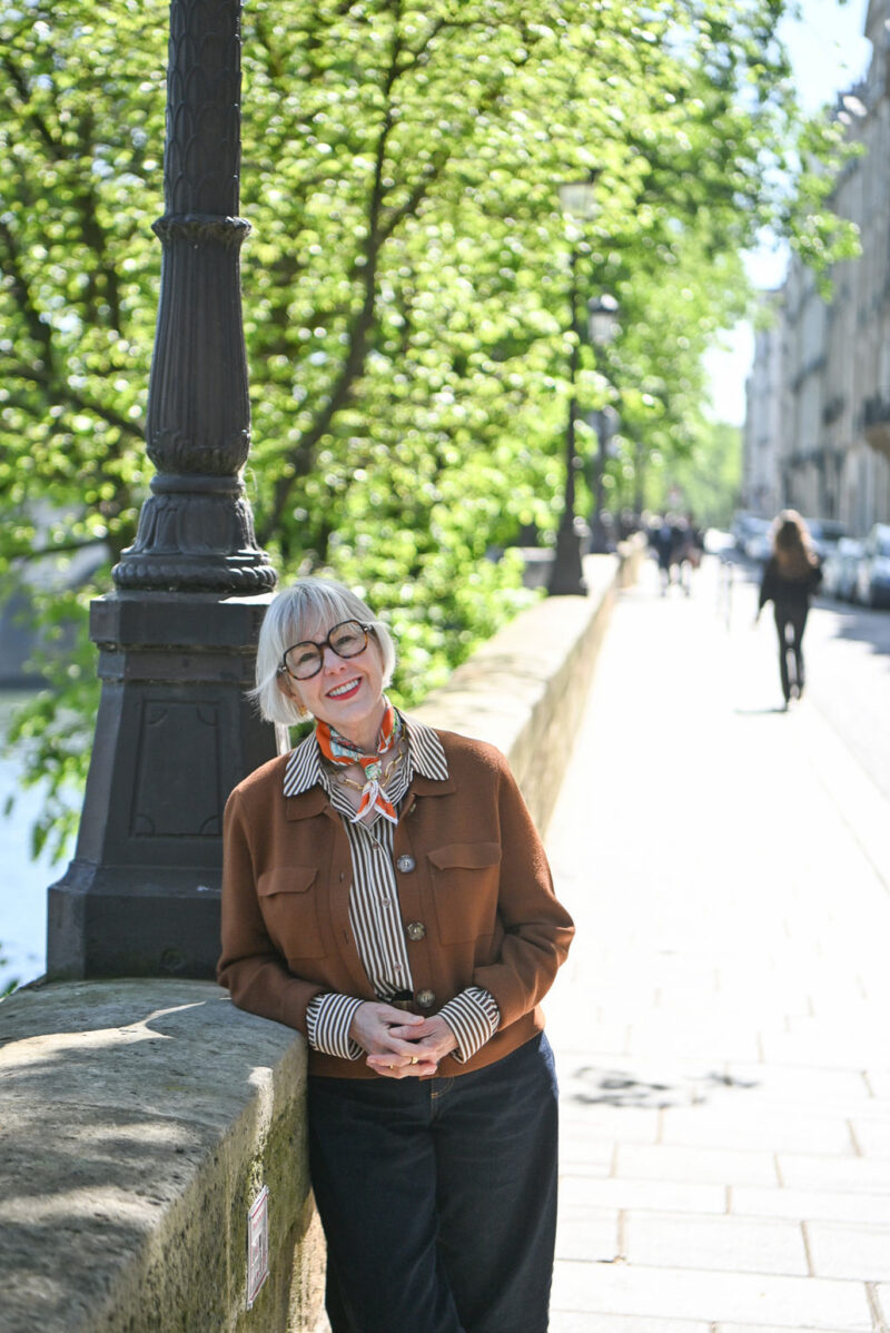 Susan B. leans against a wall on Ile St. Louis in Paris, wearing an orange scarf, striped shirt, brown jacket, and jeans.
