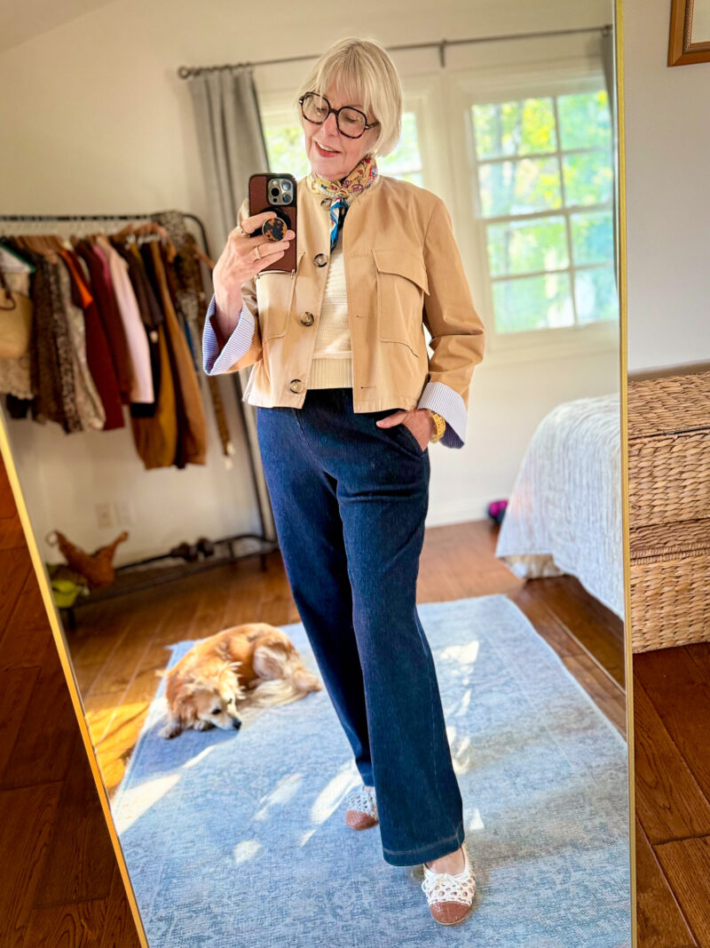 Tan utility jacket over a cream sweater and floral scarf, with dark wash straight-leg drawstring jeans and white crochet flats.
