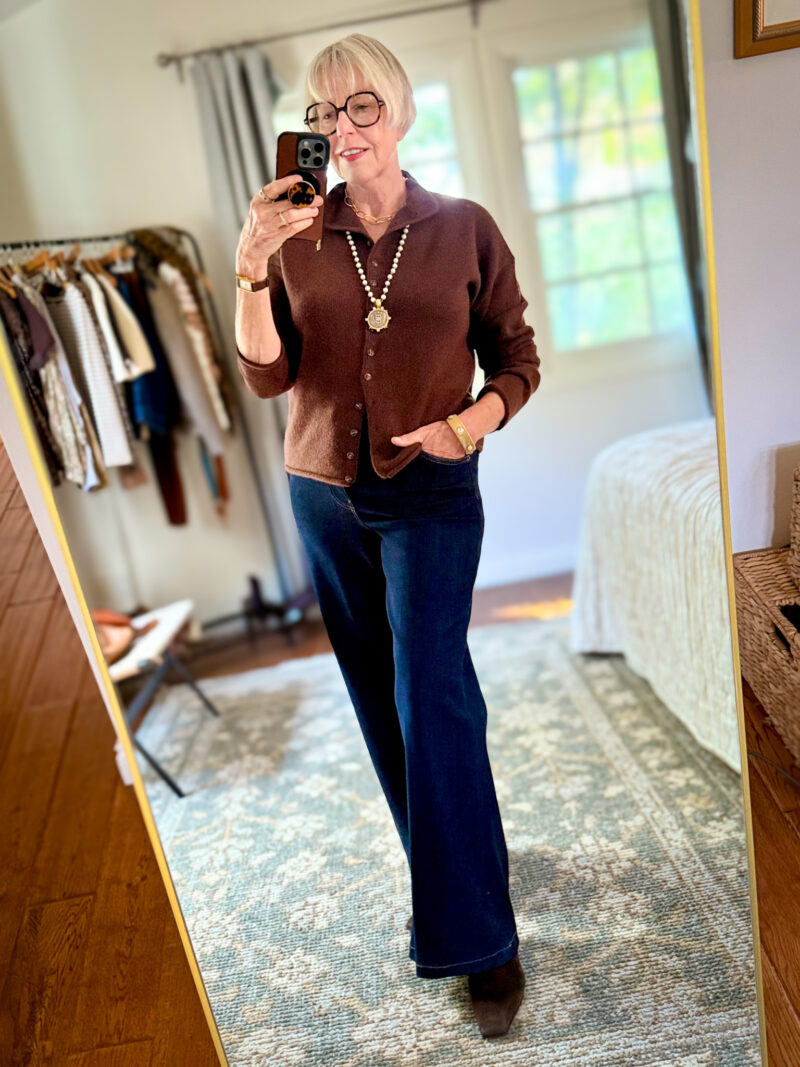 Susan B. takes a mirror selfie wearing a brown Alex Mill cashmere cardigan, French Kande pearl and medallion necklace, Ruti Palazzo dark wash jeans and brown suede boots.