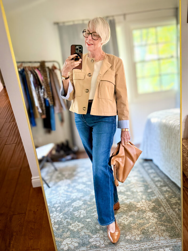 Susan wears a smooth beige utility jacket over an ivory top with dark straight-leg jeans, tan flats, and a soft leather clutch.