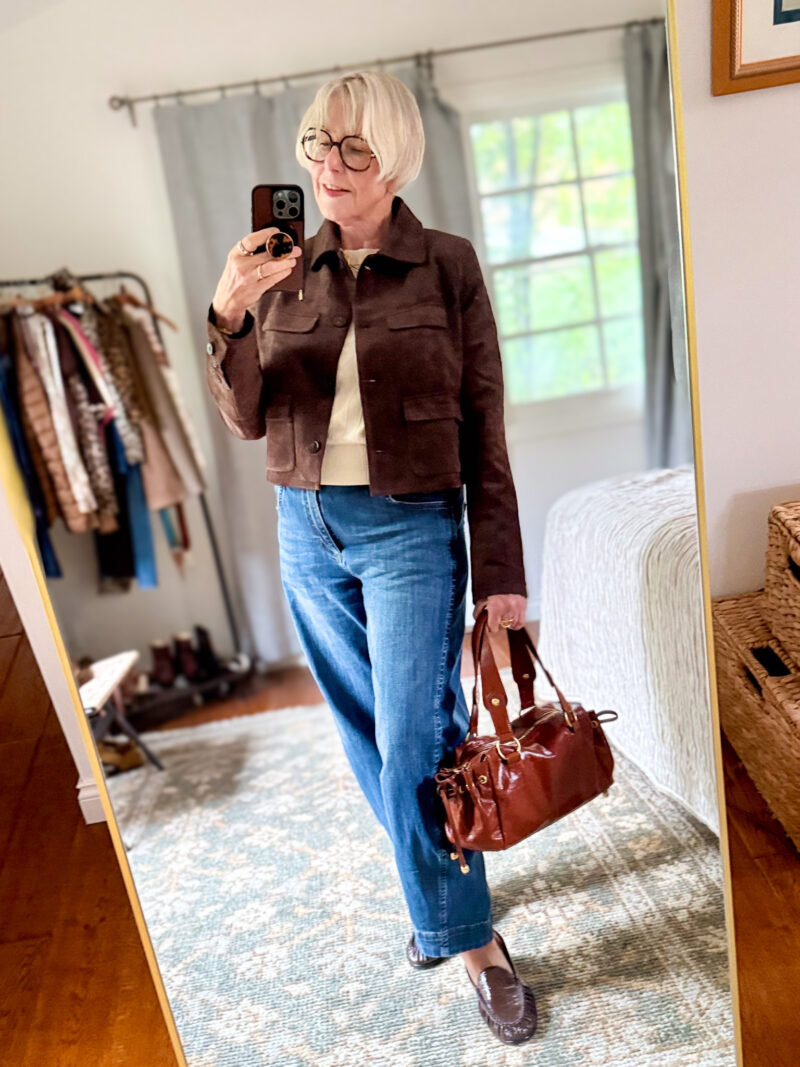 Susan wears a brown faux suede jacket over an ivory top with blue barrel jeans, brown loafers, and a glossy leather bag.