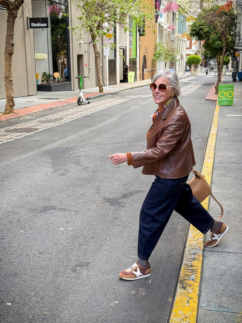 Susan B. walks across street wearing a brown leather jacket, dark wash barrel leg jeans, and brown and white sneakers.