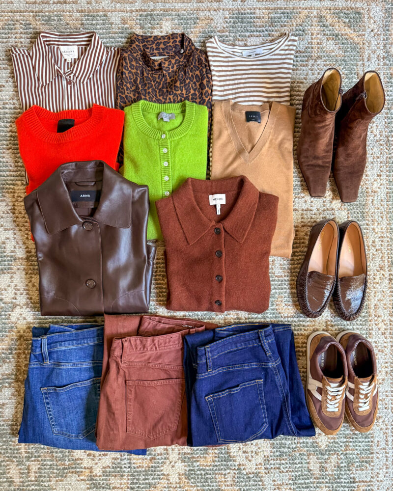 Fall Europe travel wardrobe laid out with striped and leopard print tops, colorful knits, three pairs of jeans, a brown leather jacket, boots, loafers, and sneakers.