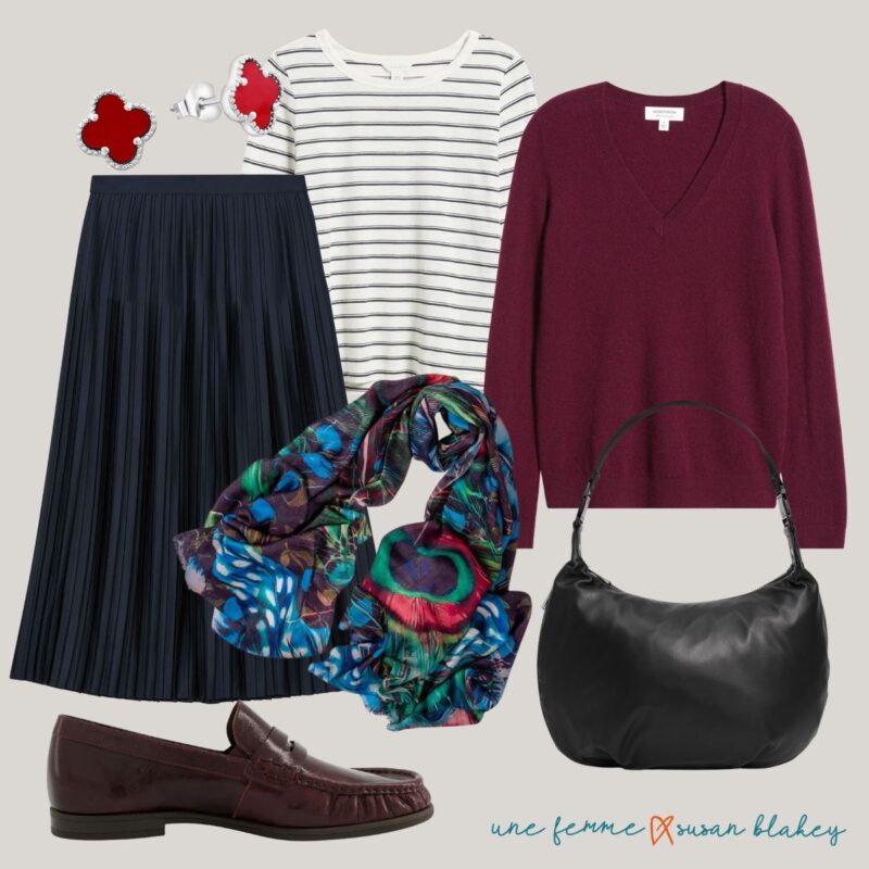 Travel outfit idea with a navy pleated skirt, silver and red earrings, striped layering tee, burgundy v-neck sweater, cashmere silk scarf in peacock feather print, burgundy loafers, and black leather bag.