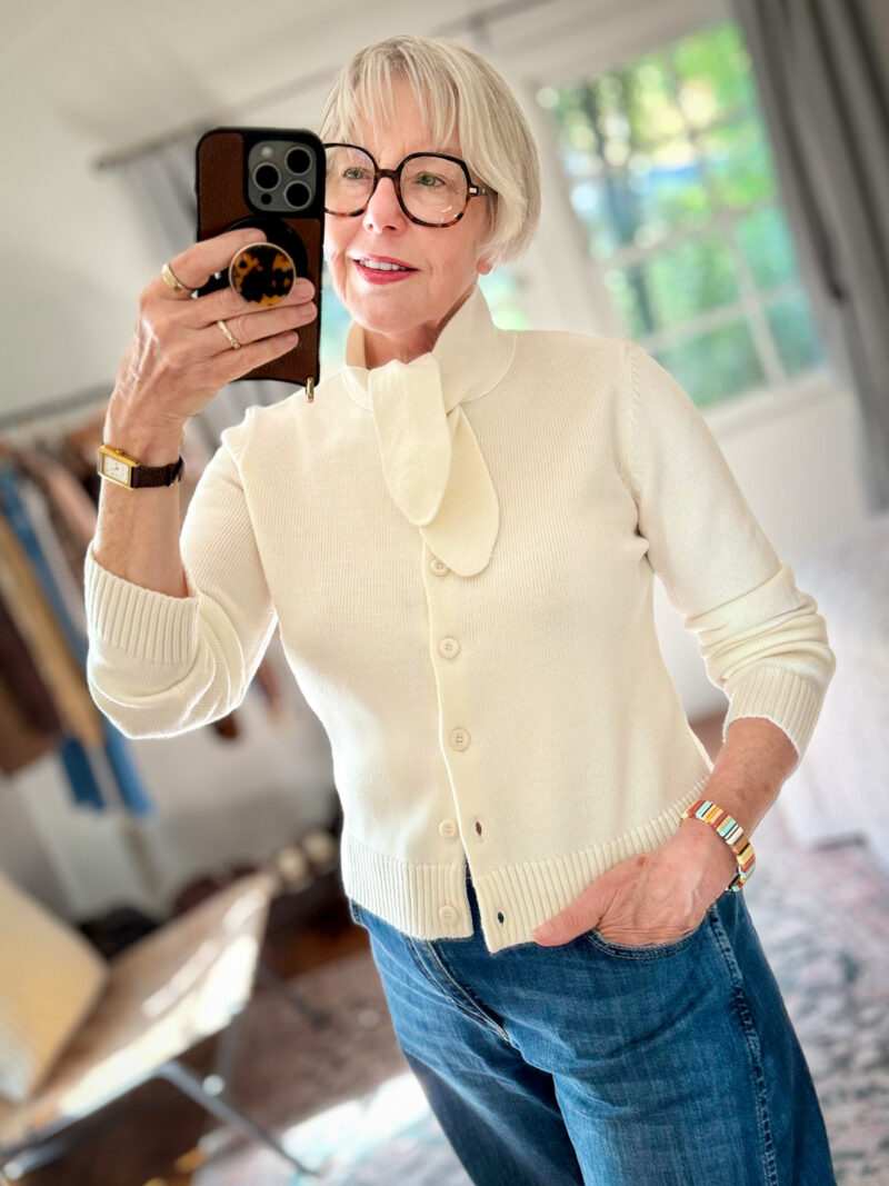 Susan B. takes a mirror selfie wearing an ecru Sezane Amelie cardigan with neck tie and medium wash jeans.
