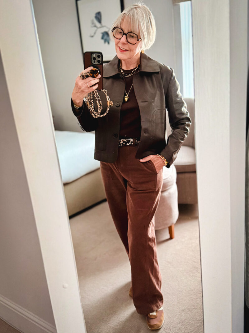 Susan B takes a mirror selfie wearing a brown leather jacket, brown sweater, acorn necklace, leopard print belt, brown pants and sneakers.