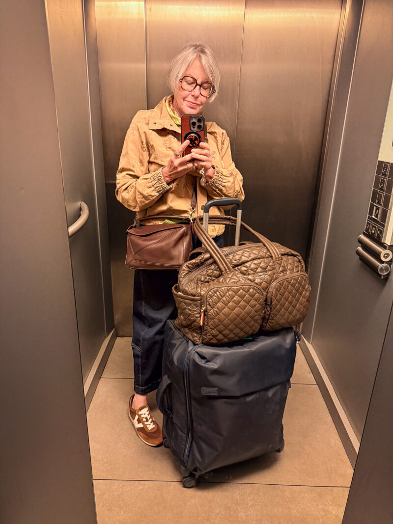 Susan taking an elevator mirror selfie with luggage, wearing a tan jacket, dark jeans, brown sneakers and a crossbody bag.