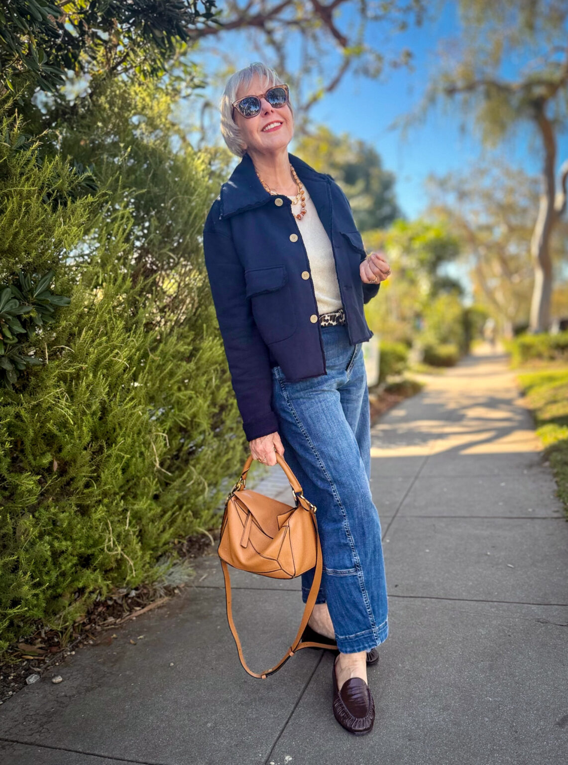 Susan wearing a navy jacket over a cream top with cropped blue jeans, a leopard-print belt, dark brown loafers, and a tan leather handbag.