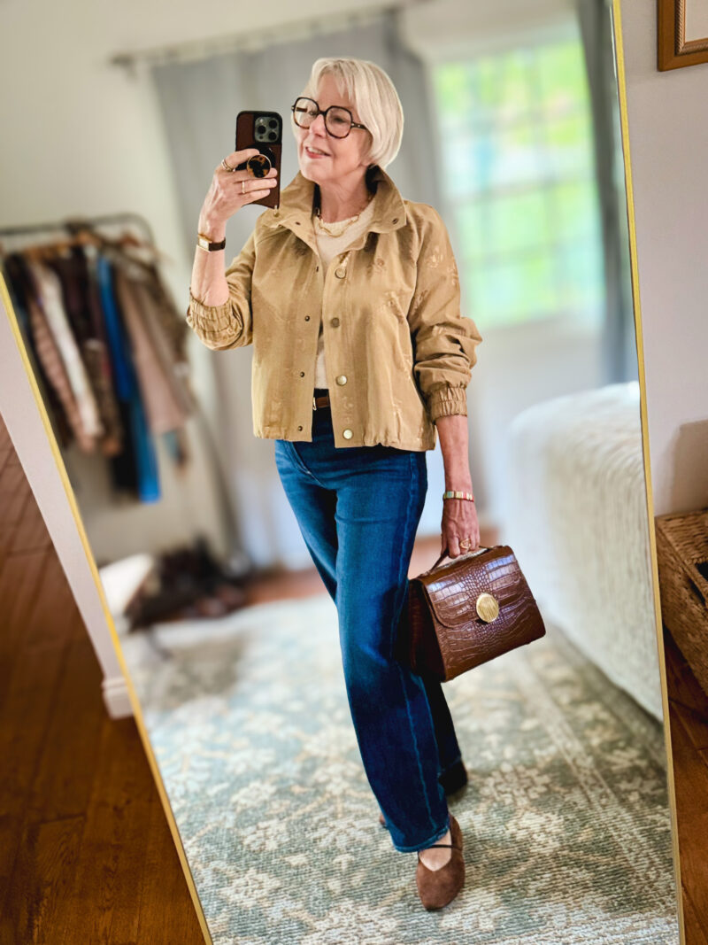 Susan wears a textured tan jacket with blue jeans and suede flats, carrying a brown croc-embossed handbag.