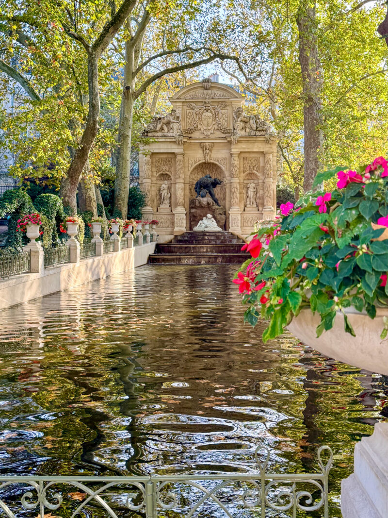 Medici Fountain in the Jardin du Luxembourg, Paris, with red and pink impatiens flowers in a planter in the foreground.