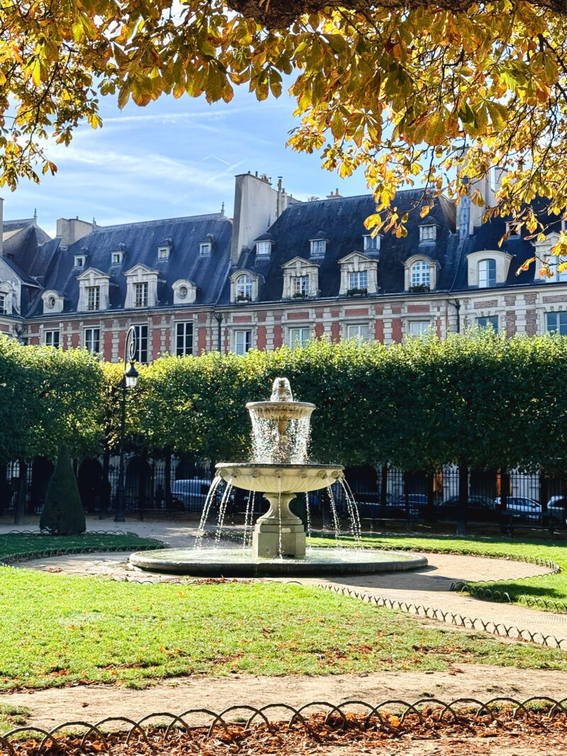 A sunlit fountain in Place des Vosges in Paris, with trees and brick buildings in the background and autumn leaves in the foreground.