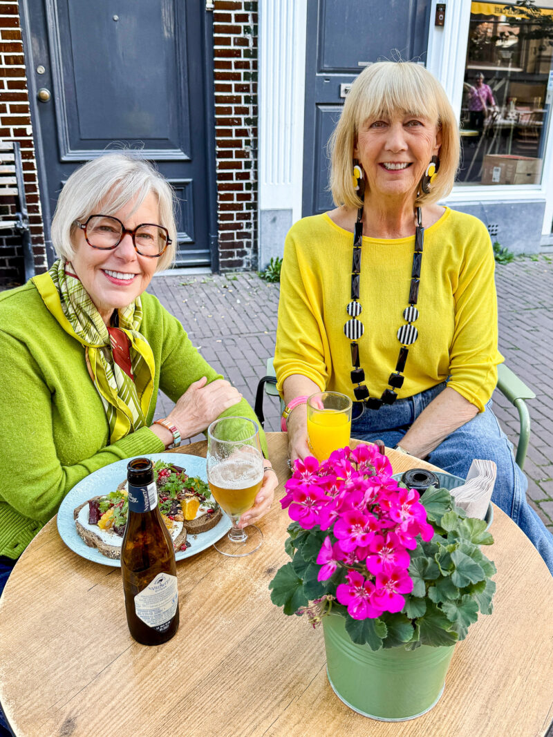 Susan and Greetje smiling over lunch at an outdoor café in Haarlem, wearing bright green and yellow sweaters.