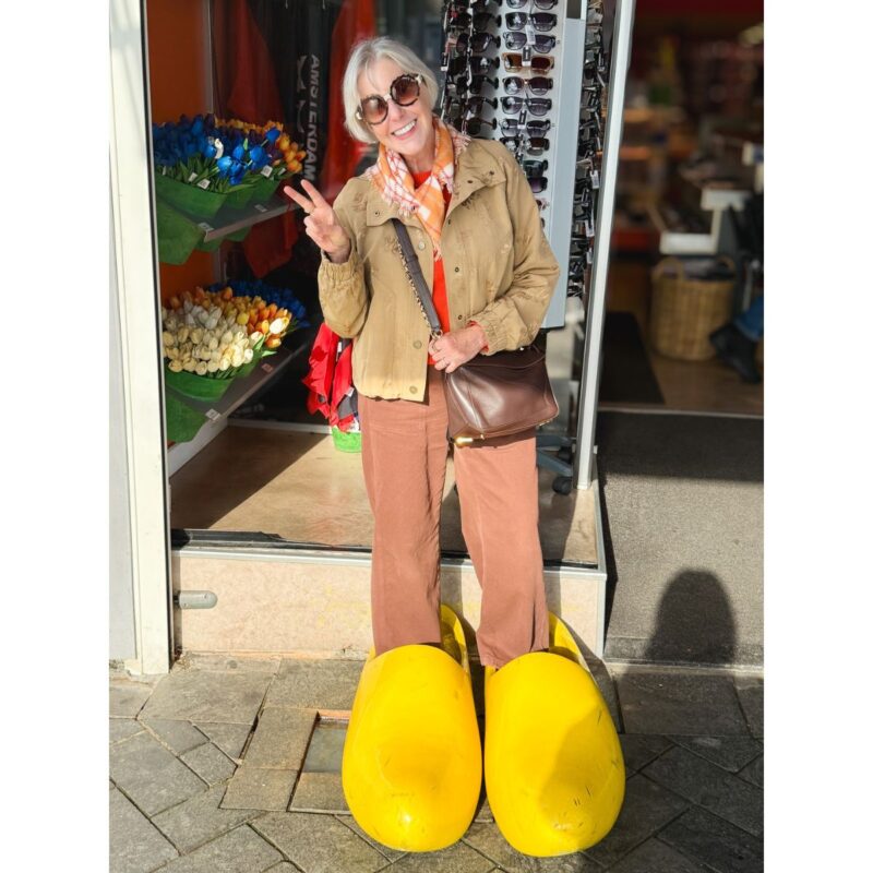 Susan wearing sunglasses and posing in oversized yellow wooden clogs outside an Amsterdam souvenir shop, holding up a peace sign.
