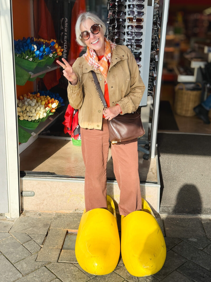 Susan wearing sunglasses and posing in oversized yellow wooden clogs outside an Amsterdam souvenir shop, holding up a peace sign.