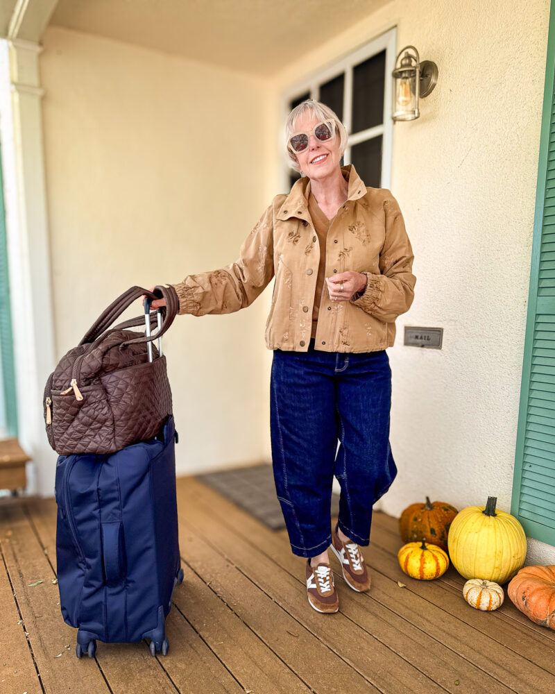 Woman wearing a Veronica Beard tan floral jacket, cropped dark blue jeans, brown sneakers and sunglasses, standing beside a navy suitcase and brown quilted bag.