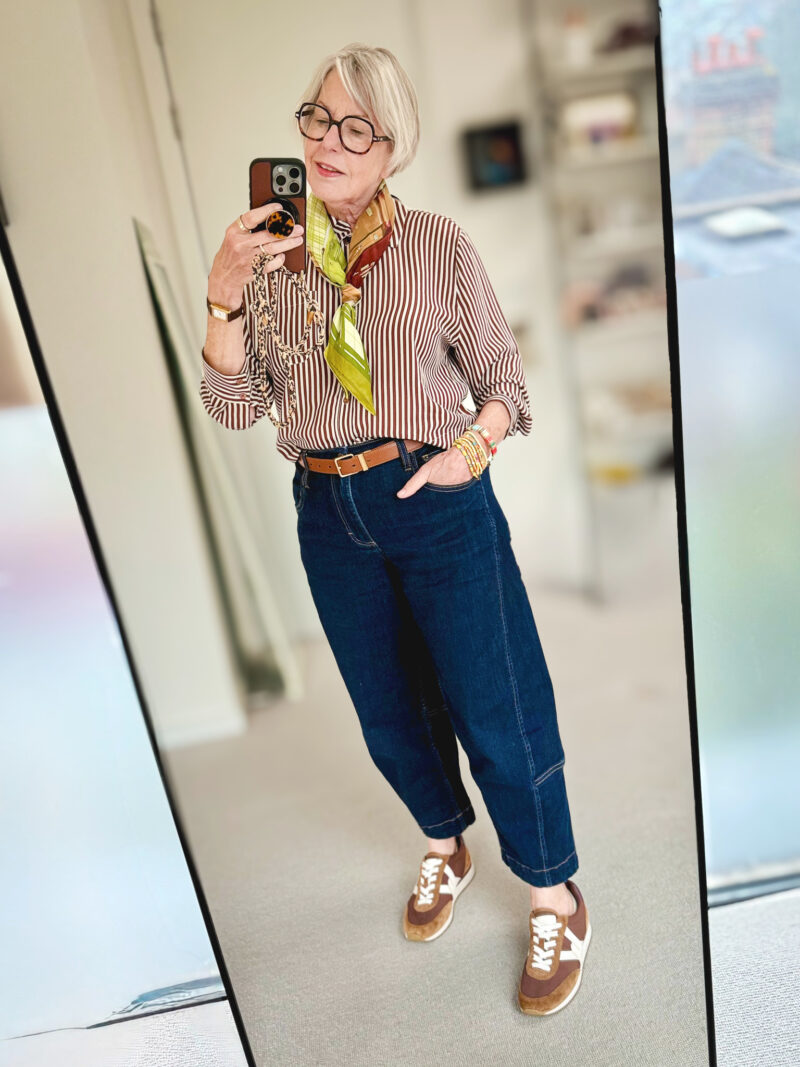 Mirror selfie of a woman wearing a brown-and-white striped shirt, cropped dark blue jeans, a tan belt, a colorful green scarf and brown sneakers.